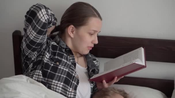 Authentieke leuke jonge vrouw of babysitter leest sprookje van boek naar kinderen voor het slapengaan. Mama zorgt voor slapende kinderen. Twee baby 's slapen in een wit bed. Moederdag, moederschap, kindertijd — Stockvideo