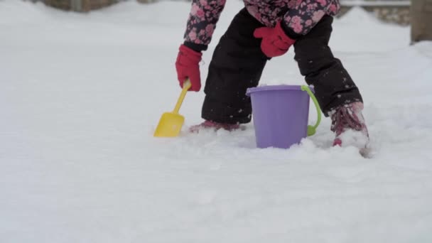 Invierno, vacaciones, juegos, conceptos familiares - plan medio de la niña de 3-4 años de edad preescolar auténtica en púrpura en el patio cubierto de nieve recoger la nieve en cubo con espátula de arena. niño en la zona nevada — Vídeos de Stock