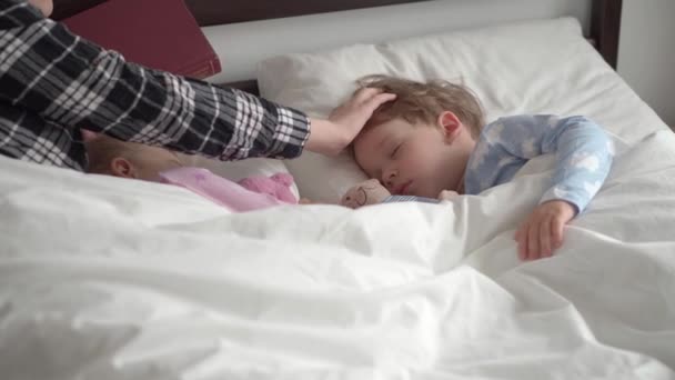 Authentieke leuke jonge vrouw of babysitter leest sprookje van boek naar kinderen voor het slapengaan. Mama zorgt voor slapende kinderen. Twee baby 's slapen in een wit bed. Moederdag, moederschap, kindertijd — Stockvideo