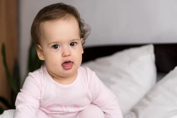 Retrato autêntico bonito caucasiano pequeno bebê gordinho menina ou menino em rosa sonolento ao acordar olhando para a câmera mostrando a língua na cama branca. Cuidados infantis, Infância, Paternidade, conceito de estilo de vida — Fotografia de Stock