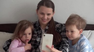 Pek çok çocuğun gerçek annesi genç bir kadın. Bebek ve kız, video konferansında akıllı telefondan konuşuyorlar. Beyaz yatakta el sallayarak gülümsüyorlar. teknoloji, annelik, kavram