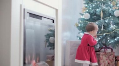 Noel Baba şapkası takan, kırmızı elbiseli, mutlu, mutlu, tombul bir kız çocuğu. Yeni yıl atmosferini kutlarken evdeki Noel ağacının yanında iyi eğlenceler. Çocukluk, bayram, kış konsepti