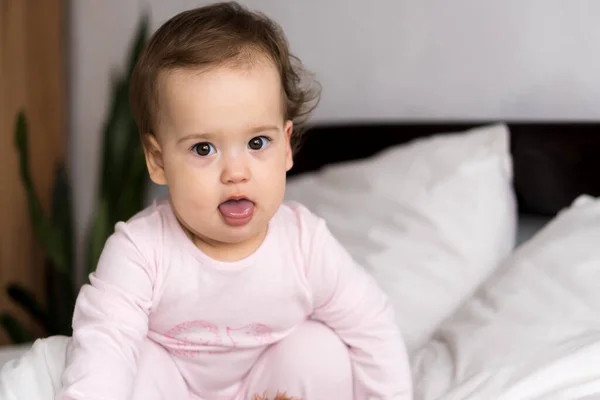 Retrato autêntico bonito caucasiano pequeno bebê gordinho menina ou menino em rosa sonolento ao acordar olhando para a câmera mostrando a língua na cama branca. Cuidados infantis, Infância, Paternidade, conceito de estilo de vida — Fotografia de Stock