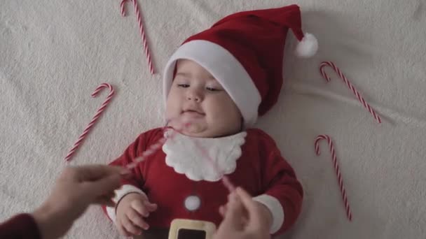 Feliz Navidad, feliz año nuevo, bebés, infancia, concepto de días festivos - primer plano sonriendo 7 meses divertido bebé recién nacido en sombrero de Santa Claus, juego de body rojo en la cama suave blanca jugar lamer dulces de Navidad — Vídeos de Stock