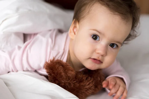 Retrato autêntico bonito caucasiano pequeno bebê gordinho menina ou menino em rosa sonolento ao acordar com ursinho olhando para a câmera na cama branca. Cuidados infantis, Infância, Paternidade, conceito de estilo de vida — Fotografia de Stock