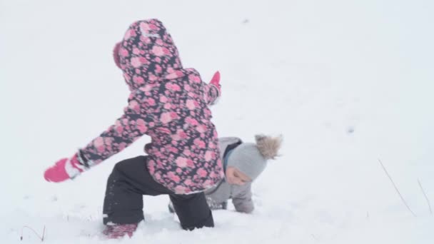 Férias, jogo, conceito de família - slo-mo autêntico Duas crianças pré-escolares felizes irmãos crianças deslizar para baixo da colina e ajudar uns aos outros subir. queda de neve na estação fria tempo no parque de inverno ao ar livre — Vídeo de Stock