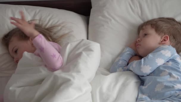 本物の白人2人の小さな兄弟就学前の赤ちゃんの女の子の男の子ピンクブルー眠いです目を覚ますとカメラを見てかわいい笑顔と白いベッドの中の優雅。育児,小児期,親,人生の概念 — ストック動画