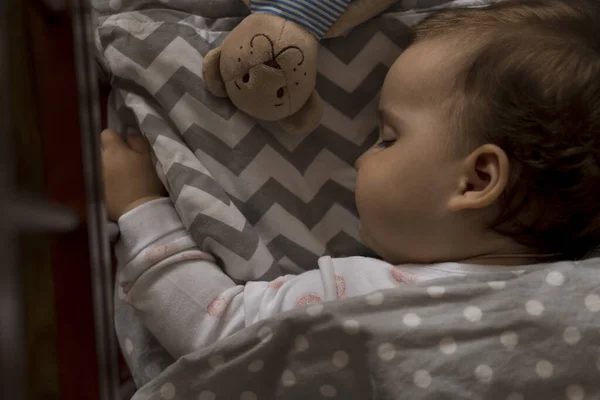 Красивая маленькая пухленькая девочка, сладко спит в серой кроватке во время обеденного отдыха в белой и розовой пижаме с плюшевым мишкой дома. Детство, отдых, комфорт, медицина, концепция здоровья — стоковое фото