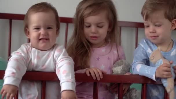 Mignon joyeux 3 petits enfants fille et garçon frères et sœurs enfants d'âge préscolaire dans la crèche de bébé gris après s'être réveillé du sommeil en pyjama avec les jambes pendantes. Enfance, loisirs, confort, médecine, concept de santé — Video