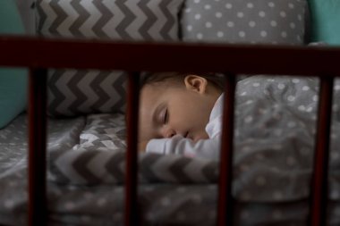 Şirin, neşeli, tombul bir kız çocuğu öğle yemeği arasında gri bebek beşiğinde tatlı tatlı uyuyor. Beyaz ve pembe pijamalarıyla evde oyuncak ayıyla. Çocukluk, rahatlık, rahatlık, ilaç, sağlık konsepti