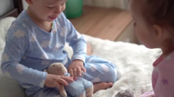 2 niños en edad preescolar sonrientes niño y niña juegan con el oso de peluche en pijama rosa azul en la cama. Los bebés gemelos se divierten, niños felices en cuarentena en casa. Amistad, familia, concepto educativo — Vídeos de Stock