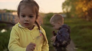 Authentic cute two little preschool sibling children girl and boy walking in park on tall grass at spring sunset. KId waving dandelion to the camera. Childhood, parenthood, family, lifestyle concept