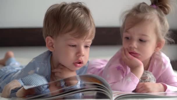 Amitié, famille, éducation concept - Deux enfants souriants d'âge préscolaire, tout-petits garçon et fille couché sur le dos en pyjama sur lit blanc. Les frères et sœurs les petits jumeaux s'amusent, les enfants heureux en quarantaine à la maison — Video