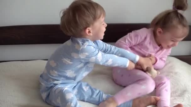 2 leende förskolebarn pojke och flicka slåss om en leksak i blå rosa pyjamas på sängen. Små tvillingar har kul, glada barn i karantän hemma. Vänskap, familj, utbildningskoncept — Stockvideo