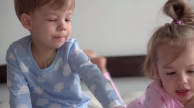Arkadaşlık, aile, eğitim konsepti. İki tane gülümseyen çocuk, beyaz yatakta pijamalarıyla uzanan çocuk ve kız çocuğu. Kardeş ikizler evde karantinada eğleniyor, mutlu çocuklar.