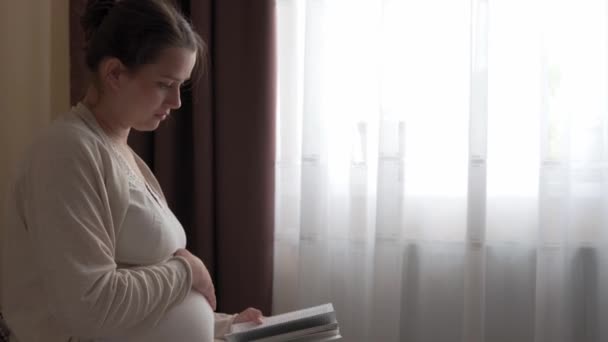 Auténtica linda mujer embarazada sintiéndose feliz en casa con vientre. Joven madre acariciando barriga y leyendo libro en la cama cerca de la ventana. Atención prenatal de maternidad, embarazo, maternidad, concepto de estilo de vida — Vídeo de stock