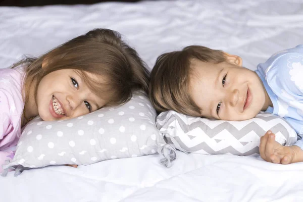Дом, комфорт, детство, уход, дружба, сладкий сон - два улыбающихся счастливый подлинный ребенок брат и сестра дети близнецы в пижаме лежат спать на кровати подушки смотреть на камеру мягкий уютный сонливое настроение — стоковое фото