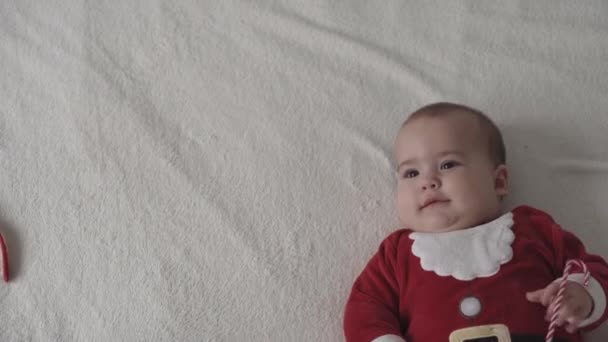 Joyeux noël, bonne année, nourrissons, enfance, concept de vacances - gros plan souriant 7 mois nouveau-né drôle dans santa claus chapeau, costume rouge jouer sur blanc doux lit jouer lécher bonbons de Noël doux — Video