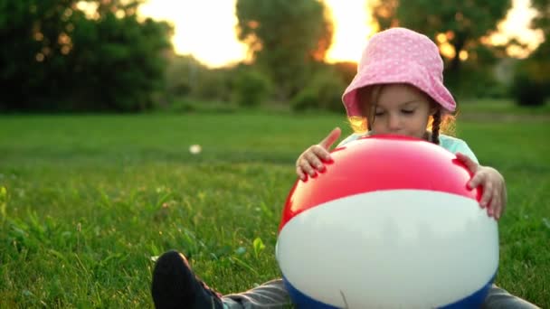 Lato, wakacje, przyroda, szczęśliwe dzieciństwo - jedno małe dziecko w przedszkolu uśmiechnięte dziecko dziewczyna w różowym kapeluszu siedzieć pozowanie patrząc na kamerę bawić się z dużym nadmuchiwane w parku na łące o zachodzie słońca świeże powietrze — Wideo stockowe