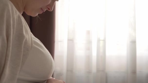 Pravá roztomilá těhotná žena se cítí šťastná doma s břichem. Mladá matka hladila bříško a četla knihu na posteli u okna. prenatální mateřská péče, těhotenství, mateřství, koncepce životního stylu — Stock video