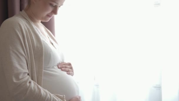 Authentique femme enceinte mignonne se sentant heureuse à la maison en prenant soin de l'enfant. Jeune femme enceinte tenant bébé dans le ventre près de la fenêtre. Maternité soins prénatals et grossesse, maternité, concept de style de vie — Video