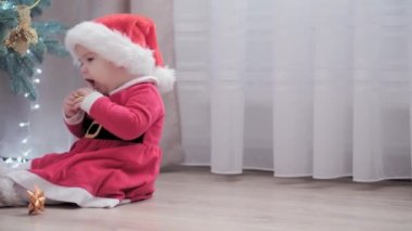 Noel Baba şapkası takan, kırmızı elbiseli, mutlu, mutlu, tombul bir kız çocuğu. Yeni yıl atmosferini kutlarken evdeki Noel ağacının yanında iyi eğlenceler. Çocukluk, bayram, kış konsepti