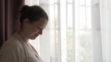 Otantik, tatlı, hamile bir kadın evde karnıyla mutlu hissediyor. Genç anne karnını okşuyor ve pencerenin yanındaki camdan su içiyor. Doğum öncesi bakım, hamilelik, annelik, yaşam tarzı konsepti