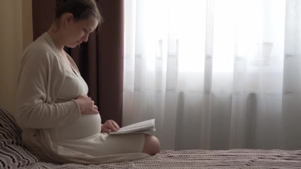 Auténtica linda mujer embarazada sintiéndose feliz en casa con vientre. Joven madre acariciando barriga y leyendo libro en la cama cerca de la ventana. Atención prenatal de maternidad, embarazo, maternidad, concepto de estilo de vida — Vídeos de Stock