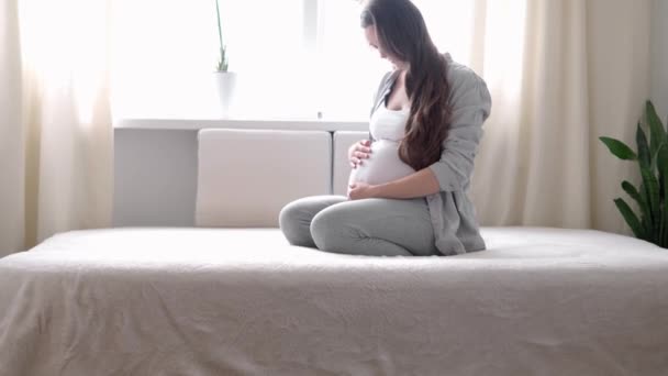Zwangerschap, moederschap, mensen, verwachting concept - gelukkige zwangere vrouw aanraken van haar buik in bed thuis zorgzame moeder streelt naakt grote buik buik met handen, op lichte kamer in zonnige zomerdag — Stockvideo