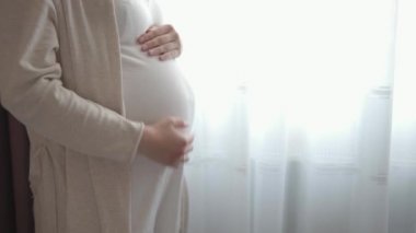 Otantik, tatlı, hamile bir kadın çocuğuna baktığı için kendini mutlu hissediyor. Gencecik bir anne karnında bebeğini pencerenin yanında tutuyor. Doğum öncesi bakım ve hamilelik, annelik, yaşam tarzı konsepti