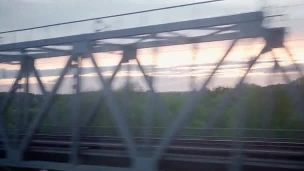 Vista da janela do trem de alta velocidade na paisagem da bela ponte metálica de campo de natureza através do rio no pôr do sol de céu crepúsculo da tarde em fundo de verão. Transporte, viagens, ferrovia, conceito de comunicação — Vídeo de Stock