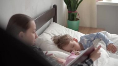 Otantik bir genç kadın ya da bebek bakıcısı yatmadan önce kitaptan çocuklara masal okur. Uyuyan çocuklara annem bakıyor. Beyaz yatakta uyuyan iki bebek. Anneler günü, annelik, çocukluk.