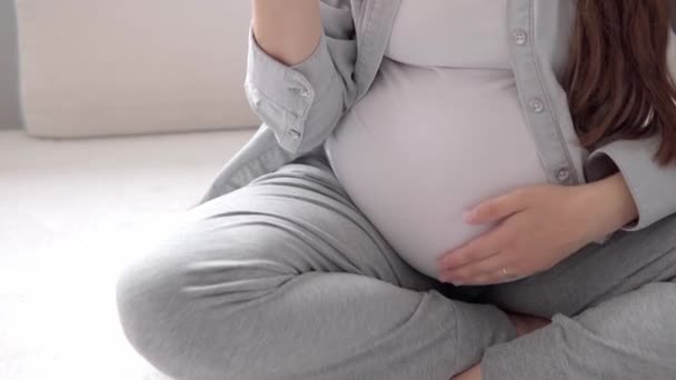 Äkta söt gravid kvinna känner sig lycklig hemma med magen. Ung mor smekte magen och åt rött på vit soffa nära fönstret. Förlossningsvård, graviditet, moderskap, livsstil — Stockvideo