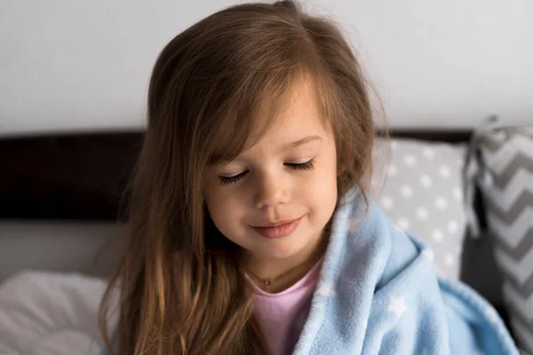 Дом, комфорт, детство, забота - одна маленькая симпатичная девочка дошкольного возраста сидит на белой серой кровати и смотрит на камеру, завернутую в теплое голубое мягкое одеяло, уютное сонное настроение в комнате внутри — стоковое фото