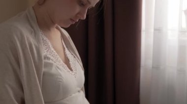 Otantik, tatlı, hamile bir kadın evde karnıyla mutlu hissediyor. Genç anne karnını okşuyor ve pencerenin yanındaki yatakta kitap okuyor. Doğum öncesi bakım, hamilelik, annelik, yaşam tarzı konsepti