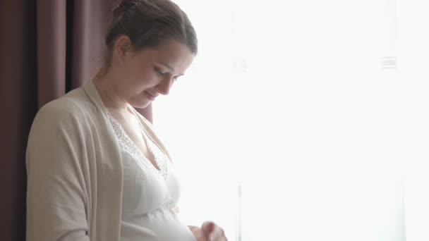 Справжня мила вагітна жінка відчуває себе щасливою вдома, піклуючись про дитину. Молода майбутня мати тримає дитину в животі біля вікна. Догляд за материнством і вагітність, материнство, концепція способу життя — стокове відео