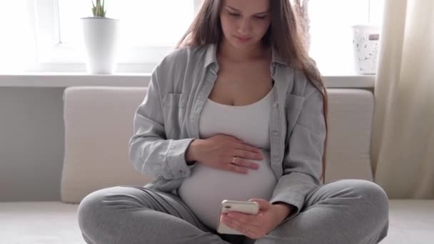 Tabletli mutlu hamile bir kadın, cep telefonuyla arkadaşlarıyla sohbet ederek kocasıyla akıllı telefonlardan iletişim kuruyor. gebelik, teknoloji, tıp — Stok video