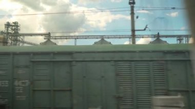 Yüksek hızlı trenin penceresinden eski paslı yük vagonlarının manzarası ve yaz arkaplanındaki güzel bulutlu mavi gökyüzünde çelik tahıl siloları. Ulaşım, seyahat, demiryolu, yol, zina kavramı