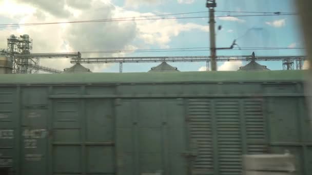 Yüksek hızlı trenin penceresinden eski paslı yük vagonlarının manzarası ve yaz arkaplanındaki güzel bulutlu mavi gökyüzünde çelik tahıl siloları. Ulaşım, seyahat, demiryolu, yol, zina kavramı — Stok video