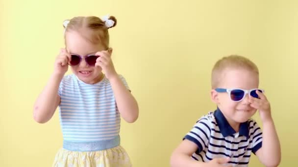 Family, friendship, childhood, summer clothing shopping. Two happy smiling toddler children sibling dance celebrate play have fun laughing. Funny kids wearing sunglasses on yellow copy space — Stock Video