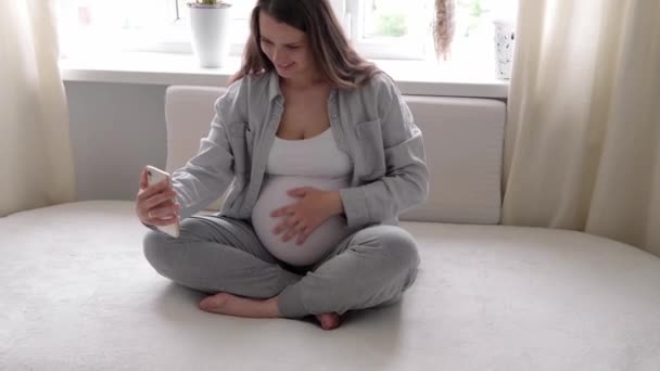 Mulher grávida feliz com tablet ter chamada de vídeo usando smartphone se comunica com o marido via chat consulta on-line com médico falar com amigos no telefone móvel. gravidez, tecnologia, medicina — Vídeo de Stock