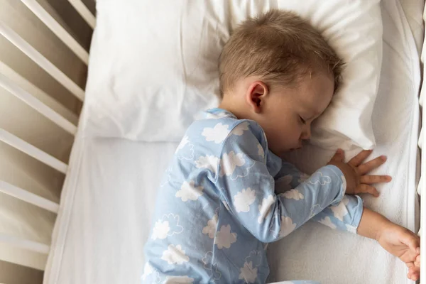 Вид сверху Милый маленький 2-3 лет дошкольный мальчик малыш спит сладко в белой кроватке во время обеденного отдыха в голубой пижаме с подушкой дома. Детство, отдых, комфорт, медицина, концепция здоровья — стоковое фото