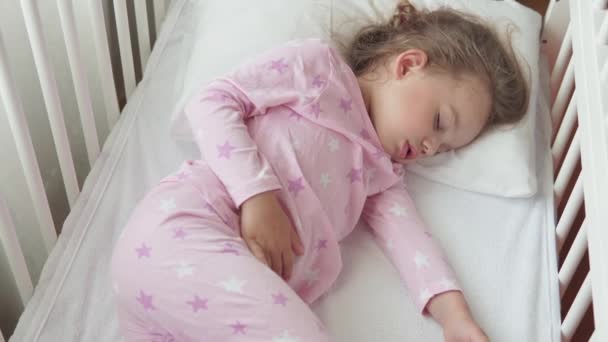 Fechar autêntico bonito caucasiano pequena menina pré-escolar dormir docemente em berço branco no pijama no quarto durante a hora de almoço. O miúdo está deitado na cama perto da janela. Infância, medicina, conceito de saúde — Vídeo de Stock
