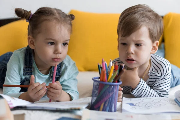 Schick lächelnd zeichnen Kleinkinder mit Bleistiften, die ihnen auf dem Bauch im gelben Bett liegen. kleine süße Leser haben Spaß, glückliches Kind in Quarantäne zu Hause. Freundschaft, Familie, Bildung — Stockfoto