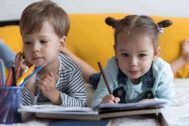 Zeki, gülümseyen, küçük bir çocuk çocuk ve kız sarı yatakta göbeklerinde kalemlerle resim yapıyorlar. Küçük sevimli okuyucu evde karantinada eğleniyor, mutlu çocuk. Arkadaşlık, aile, eğitim.