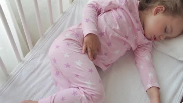 Закрыть настоящий милый кавказский маленький дошкольный ребенок девочка спать сладко в белой кроватке на пижаме в спальне во время обеда. Ребенок отдыхает лежать в постели у окна. Детство, медицина, концепция здоровья — стоковое видео