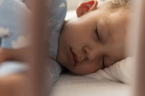 Schattig vrolijk klein 2-3 jaar kleuter kindje jongen slapen zoet in wit wieg tijdens de lunch rusttijd in blauwe pyjama met kussen thuis. Jeugd, vrije tijd, comfort, geneeskunde, gezondheidsconcept — Stockfoto