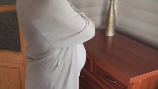 Mutlu hamile kadın pencerenin yanındaki aynanın önünde uzun saçlarını tarıyor. Şefkatli anne karnını okşar ve elleri ışık kaynağı odasındadır. hamilelik, annelik, beklenti konsepti — Stok video