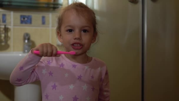ポートレート幸せなかわいい未就学児の女の子でパジャマブラッシング歯と浴室でカメラを見ています。子供の毎日の医療ルーチン。白人の少女が家で洗濯している。ライフスタイル、子供時代のコンセプト — ストック動画