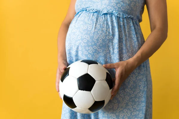 Motherhood, femininity, football, sport, dairy, hot summer. croped unrecognizable pregnant young pretty woman in floral blue dress holds soccer white and black ball rubs tummy on yellow background Stock Picture